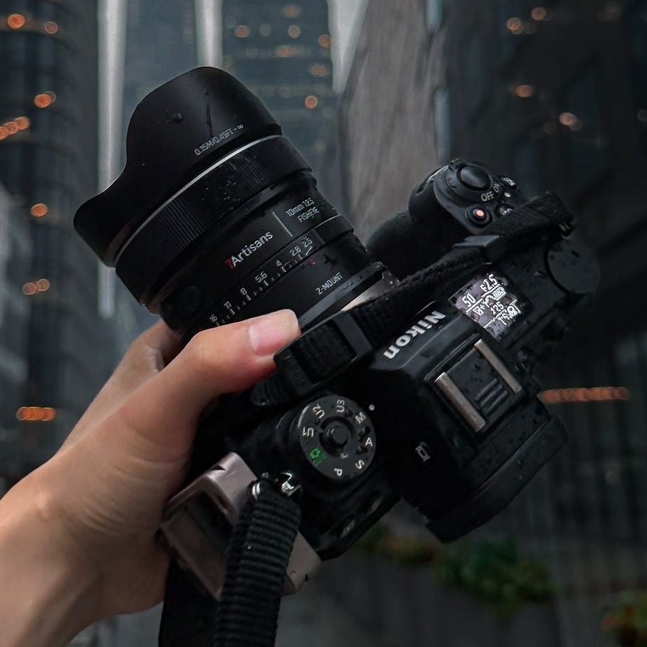 A hand holding a black Nikon mirrorless camera with a 7artisans 10mm attached, photographed from a low angle in a city street with tall blurred buildings in the background.