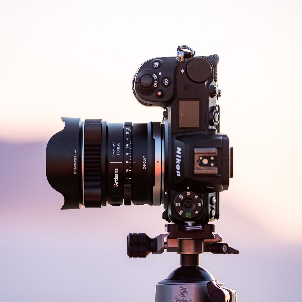 A black Nikon mirrorless camera with a 7artisans 10mm wide-angle lens attached, mounted on a tripod against a soft pastel sunset sky background.